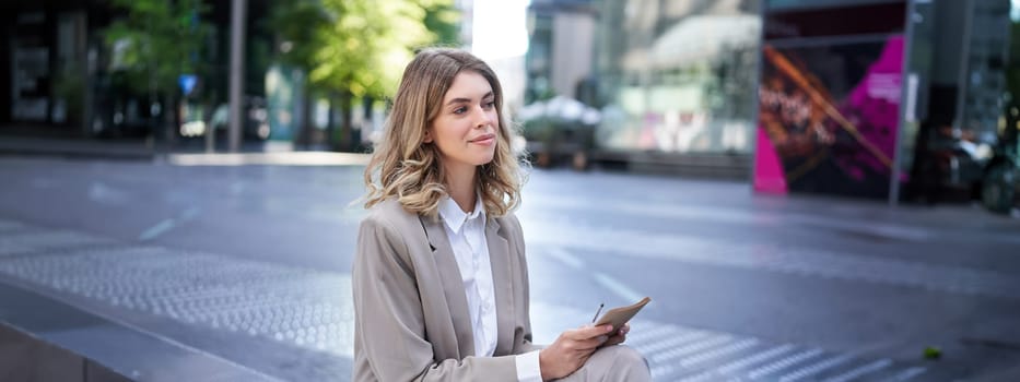 Young businesswoman preparing for speech, interview in company, writing down notes while sitting outside in city centre.