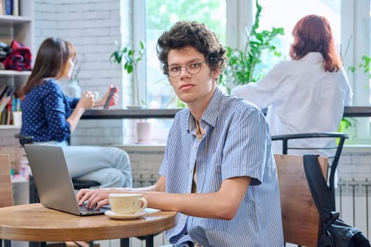 Handsome young curly guy college student with laptop cup of coffee, sitting at table in coffee shop, looking at camera. Internet online technology for leisure, learning, chat, blog, youth lifestyle