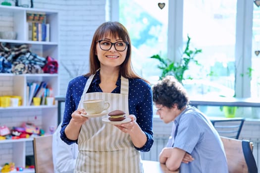 Portrait of successful female coffee shop owner holding fresh cup of coffee and cake on plate. Smiling middle aged woman waiter bakery worker in apron. Food service occupation, staff, job, work
