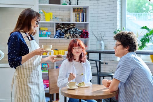 Woman in an apron with cup of coffee and plate of cake serving young people sitting at table in coffee shop, cafeteria, bakery. Food service occupation, small business, staff, job work, people concept