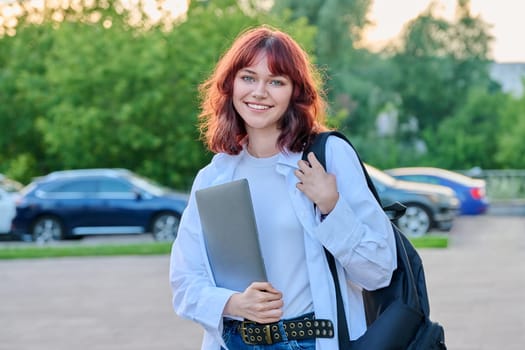 Outdoor portrait of young smiling female college student with backpack. Attractive girl with red hair holding laptop, looking at camera. 18-20 years students, youth, education, lifestyle concept