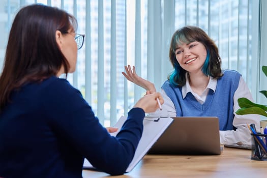 Middle aged woman teacher mentor talking helping testing female teenage student sitting in office in college building. Education, learning, contact teacher student concept