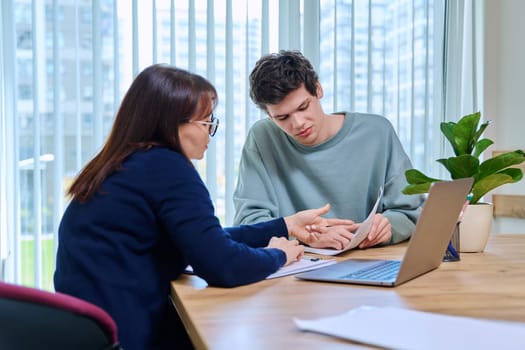 Middle aged woman teacher mentor talking helping testing male teenage student sitting in office in college building. Education, learning, contact teacher student concept