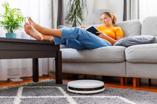Robotic vacuum cleaner cleaning a room while a woman relaxing, reading book on the sofa. Innovative technologies for cleaning