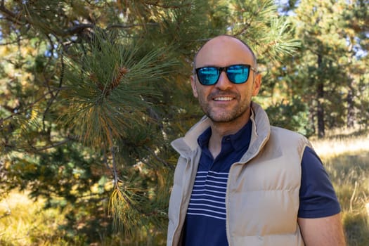 Portrait Smiling 40 yo Man in Sunglasses, Fir Tree, Forest On Background. Nature in Fall Leaves Season. Horizontal Plane. Active Lifestyle. Male Trekking or Hiking. Copy Space. High quality photo