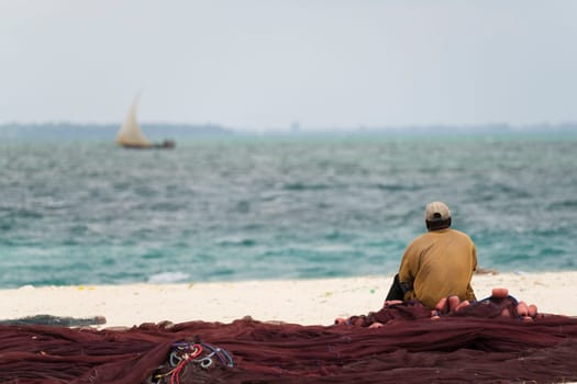 African fisherman sitting on fishing nets looks out to sea, overcast day, Zanzibar , Tanzania.