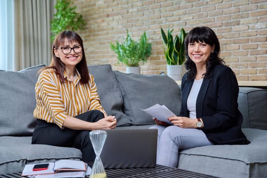 Two mature business women colleagues working together in office sitting on couch in lounge. Smiling posing females looking at camera holding business papers contracts. 40s 50s business people work job