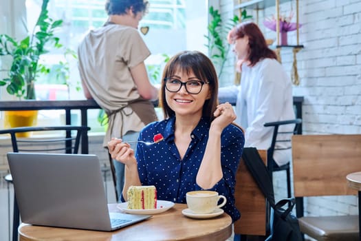 Middle aged woman in bakery cafeteria with cup of coffee and dessert cake, sitting at table, with laptop, happy smiling female looking at camera. Lunch break, food, lifestyle, mature 40s people concept