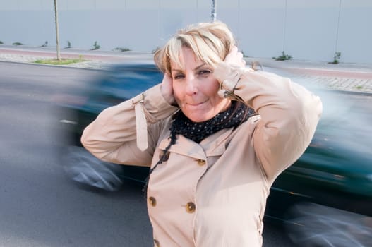 Head and shoulders shot of a mature woman in a trench coat on the side of the road with blurred cars passing by in the background,covering her ears with her palms.