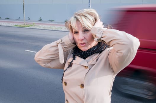 Head and shoulders shot of a mature woman in a trench coat on the side of the road with blurred cars passing by in the background,covering her ears with her palms.