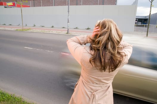 Head-and-shoulders shot of a mature woman from behind in a trench coat on the side of the road with blurred cars passing by in the background,covering her ears with her palms.