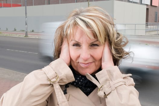 Head and shoulders shot of a mature woman in a trench coat on the side of the road with blurred cars passing by in the background,covering her ears with her palms.