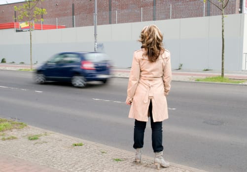 Full length shot of a mature woman in a trench coat from behind on the side of the road with blurred passing cars in the background