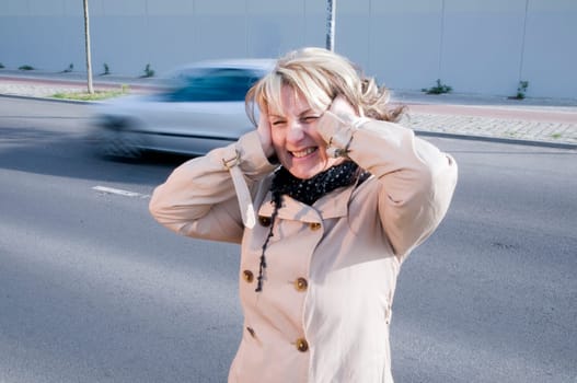 Head and shoulders shot of a mature woman in a trench coat on the side of the road with blurred cars passing by in the background,covering her ears with her palms.