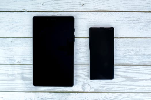 Black tablet and smartphone with turned-off screens placed side by side on a white wooden surface. Top-down view.