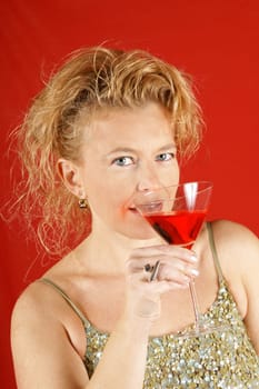 Portrait of a beautiful blond woman with blue eyes, wearing a green sequins top, holding a red drink, over a red background. Soft focus.
