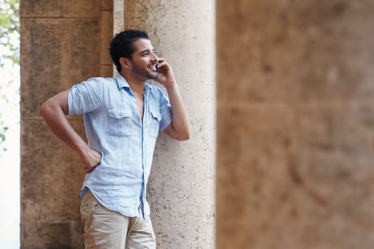 mid adult hispanic man talking on mobile phone and leaning on columns outdoors. Horizontal shape, side view, copy space