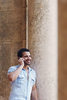 mid adult hispanic man talking on mobile phone and leaning on columns outdoors. Verticl shape, side view, copy space