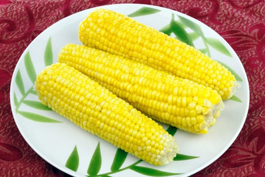 Three bright, buttered yellow vegetable ears on a bamboo design plate on red place mat.