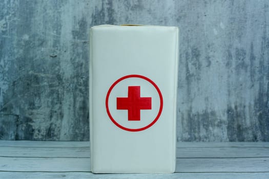 White first aid kit box featuring a red cross symbol, placed on a wooden surface against a textured gray background.