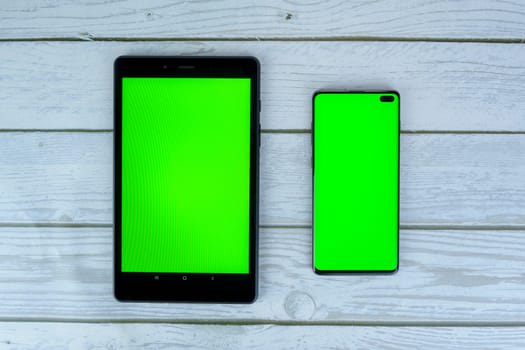 Black tablet and smartphone with blank green screens lying on a white wooden surface, ready for mockup or design presentation.