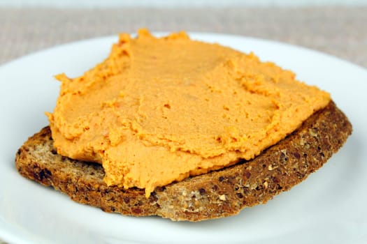 Whole grain bread is toasted, topped with red pepper hummus, served on a white plate.