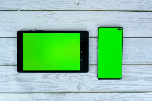 Black tablet and smartphone with blank green screens lying on a white wooden surface, perfect for mockups, design presentations, or advertising purposes.