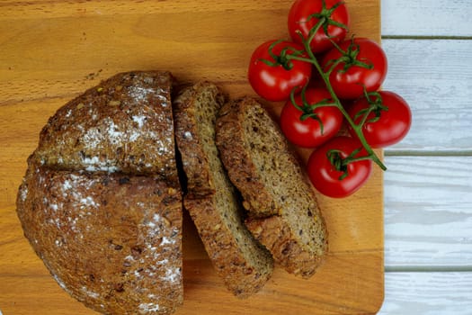 Freshly baked whole grain bread with seeds and ripe red vine tomatoes on a wooden cutting board, viewed from above on a rustic white wooden table.