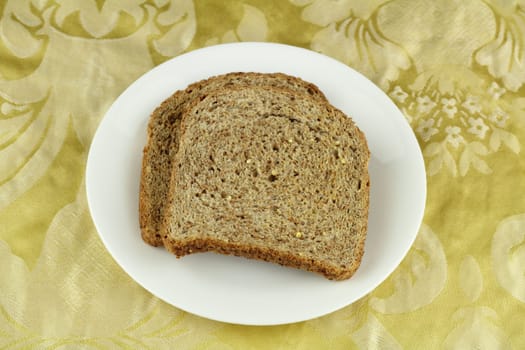 Two crisp whole sprouted grain and legume bread slices on a round white dish above a buttery yellow floral cloth placemat.
