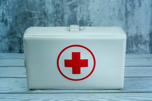 Vintage white first aid kit box with a red cross symbol, placed on a wooden surface against a gray textured background. Ideal for medical and emergency themes.