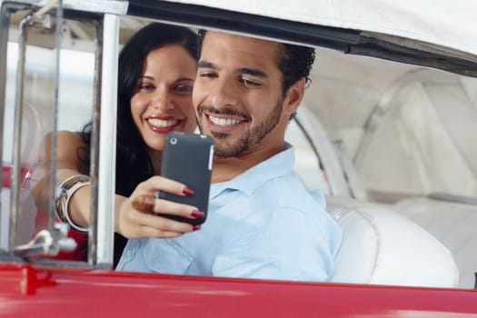 happy young man and woman smiling while taking snapshot with cell phone camera from red vintage convertible car