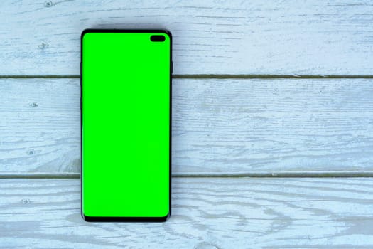 Black smartphone with blank green screen lying on a white wooden surface, ideal for mockups, design presentations, or advertising purposes.