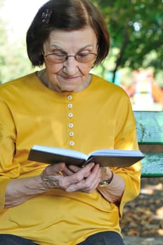 Senior woman sitting on bench reading book outdoors in park