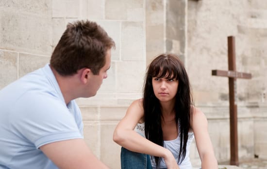 Young couple sitting outdoors ahead of church (cross in background) having relationship problems