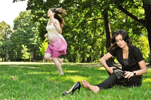 One woman running barefoot in park and second is taking her shoes off