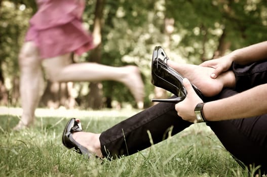 Torso of one woman running barefoot in park and second is taking her shoes off