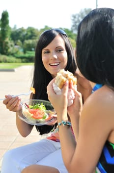 Two friends - smiling young women eating together outdoors on sunny day