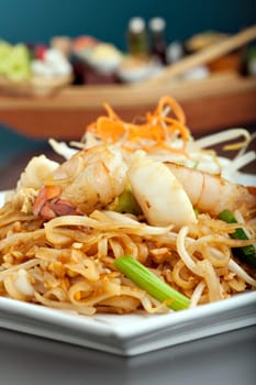 Seafood pad Thai dish of fried rice noodles on a square white plate with chopsticks and grated carrot garnish. Shallow depth of field.