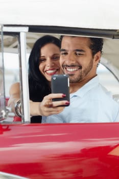 happy young man and woman smiling while taking snapshot with cell phone camera from red vintage convertible car