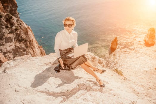 Business woman on nature in white shirt and black skirt. She works with an iPad in the open air with a beautiful view of the sea. The concept of remote work