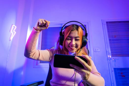 Winning. Happy Gamer playing video game online with smartphone with neon lights she raises hands to wins celebrating, Asian woman wearing gaming headphones feeling excited at home, E-Sport concept