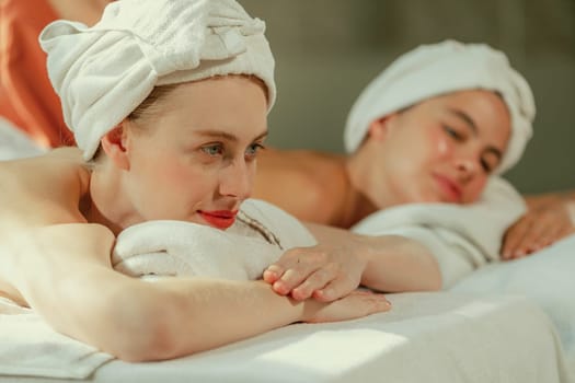 A couple of beautiful young woman with white towel lie on comfortable spa bed during receive relaxing body massage by professional masseur surrounded at spa salon. Gray background. Tranquility.