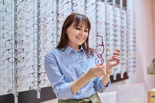 Middle-aged woman in optician's store buying, choosing glasses frames, near display case with vision eyeglasses spectacles. Optics, ophthalmology, age vision problems