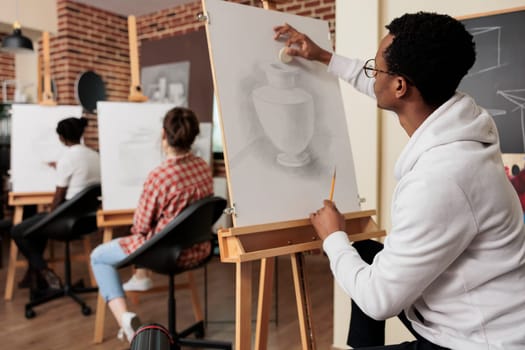 African American guy sitting at easel learning drawing techniques, sketching vase on canvas while attending creative group art class with friends. Creativity and leisure concept