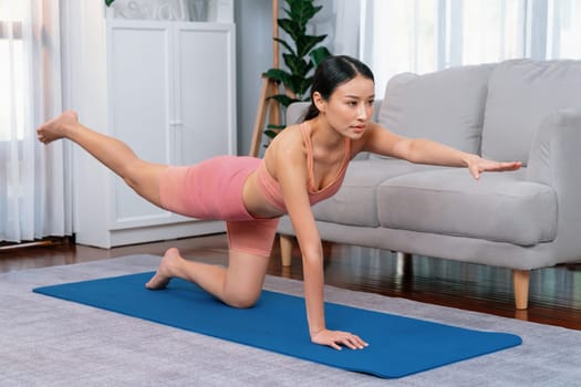 Asian woman in sportswear doing yoga exercise on fitness mat as her home workout training routine. Healthy body care and calm meditation in yoga lifestyle with comfortable and relaxation. Vigorous