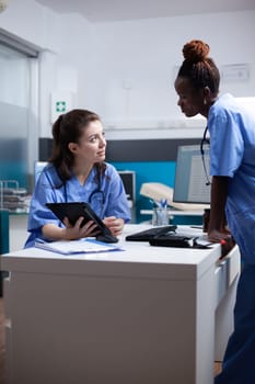 Nurse colleagues in happy relaxed hospital workspace working on tablet in modern professional medical office. Diverse healthcare specialists coworkers in modern clinic using technology