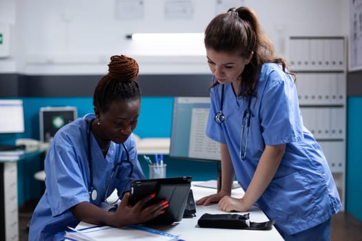 Nurse showing medical data to young collegue on a digital tablet screen in modern professional clinic office. Diverse clean modern hospital workplace equipped with technology