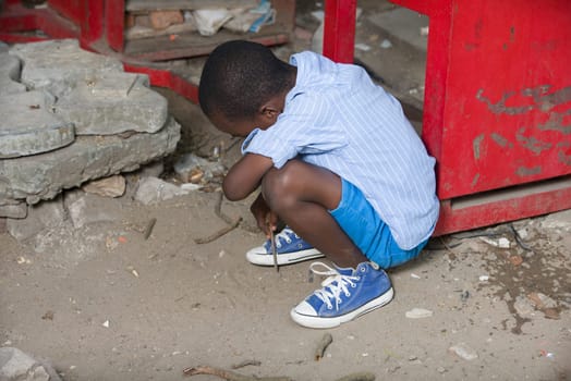 young boy in striped shirt crouched outdoors writing on the ground with a piece of wood.