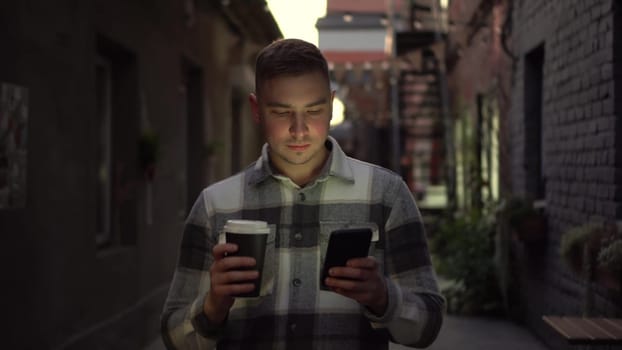 A young man walks along a narrow street late at night chatting on his smartphone and drinking coffee. A man in a warm shirt walks with a phone in his hands and drinks a hot drink. 4k