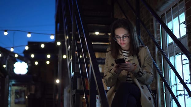 A young woman sits on a fire escape and texts on a smartphone. A girl with glasses on a narrow street late at night with a phone in her hands. 4k